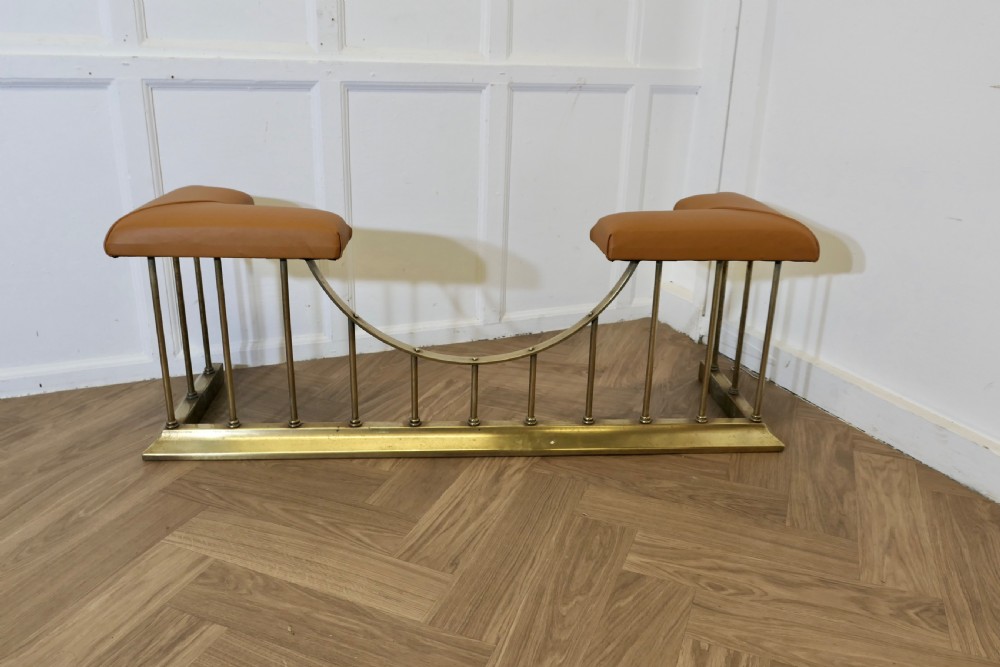 A brass club fender featuring twin tan leather upholstered corner seats supported by vertical brass spindles on a moulded base.