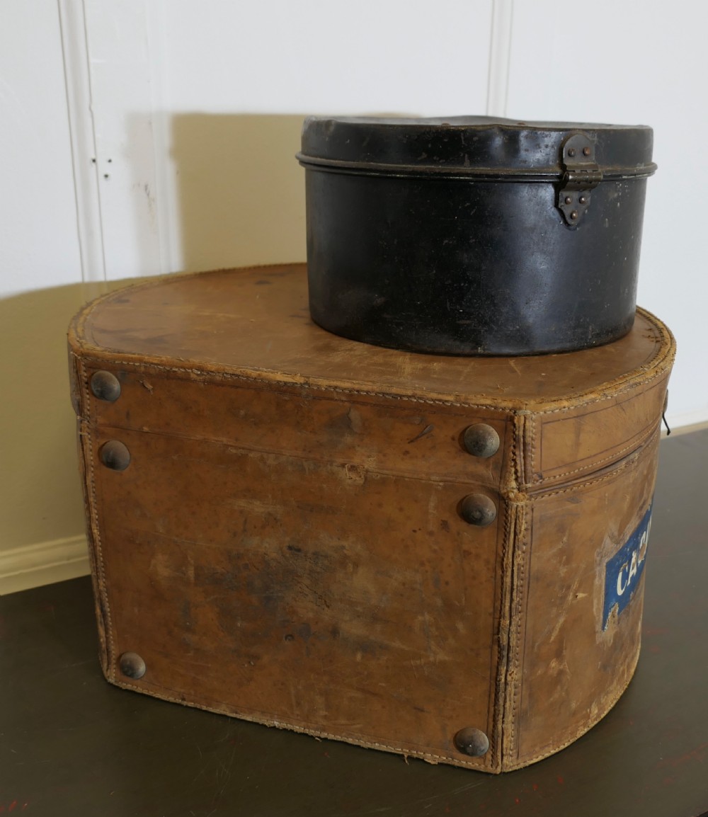 A nineteenth-century tan leather hat box featuring travel stickers and a leather handle, topped with a smaller black metal hat case. View III