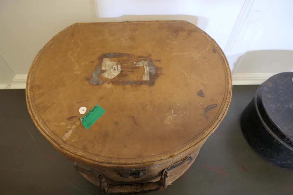 A nineteenth-century tan leather hat box featuring travel stickers and a leather handle, topped with a smaller black metal hat case. View V