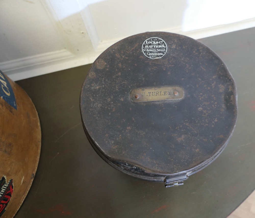 A nineteenth-century tan leather hat box featuring travel stickers and a leather handle, topped with a smaller black metal hat case. View VI