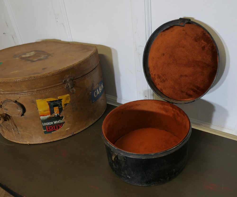 A nineteenth-century tan leather hat box featuring travel stickers and a leather handle, topped with a smaller black metal hat case. View X