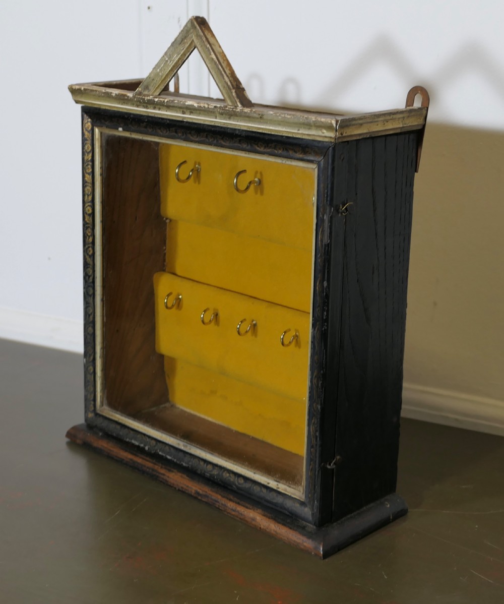 An ebonised wooden key cabinet featuring a glazed front door, triangular pediment, mustard velvet lining, and two rows of brass hooks inside. View III