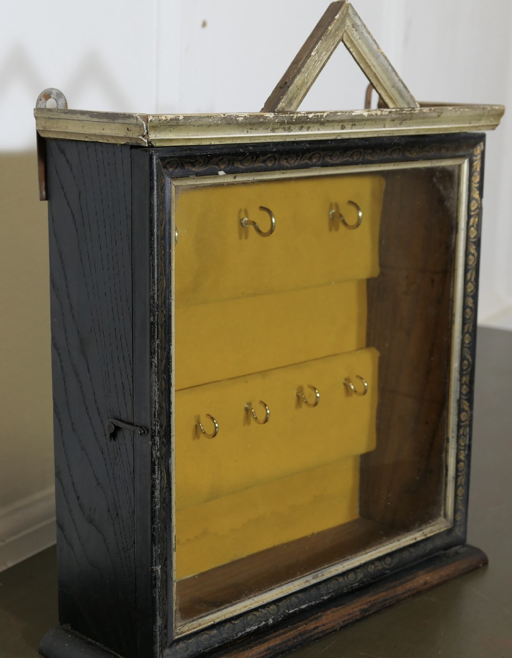 An ebonised wooden key cabinet featuring a glazed front door, triangular pediment, mustard velvet lining, and two rows of brass hooks inside. View IV