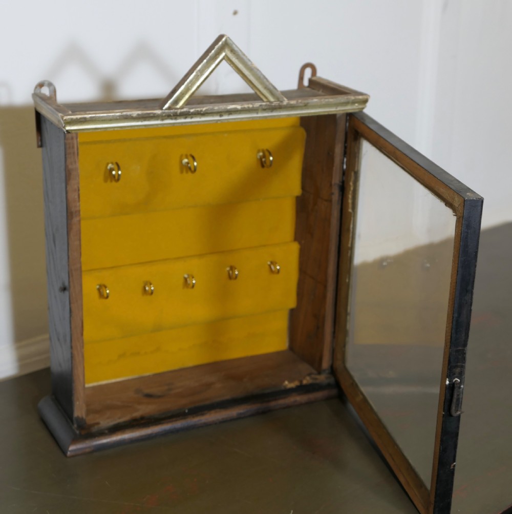 An ebonised wooden key cabinet featuring a glazed front door, triangular pediment, mustard velvet lining, and two rows of brass hooks inside. View V