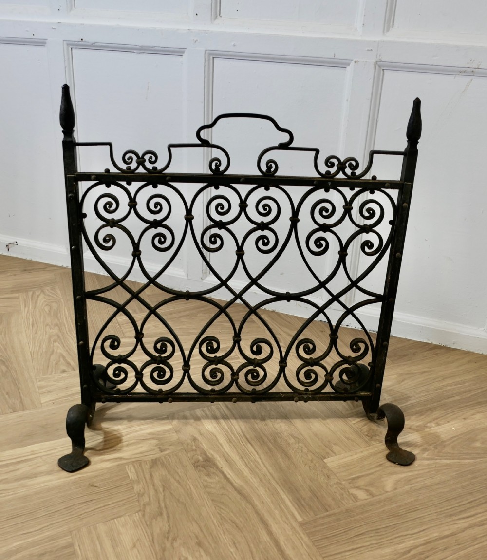 A black wrought iron fire screen featuring intricate scrollwork lattice, pointed side finials, and an integrated top handle. View VI