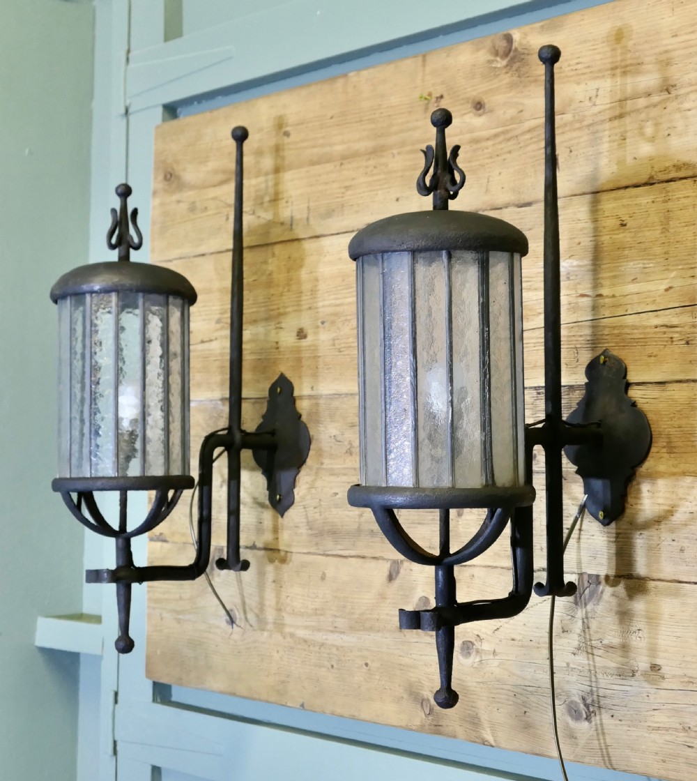 A pair of large black wrought iron wall lanterns featuring cylindrical glass shades with vertical metal bars and decorative finials at the top and bottom. View X