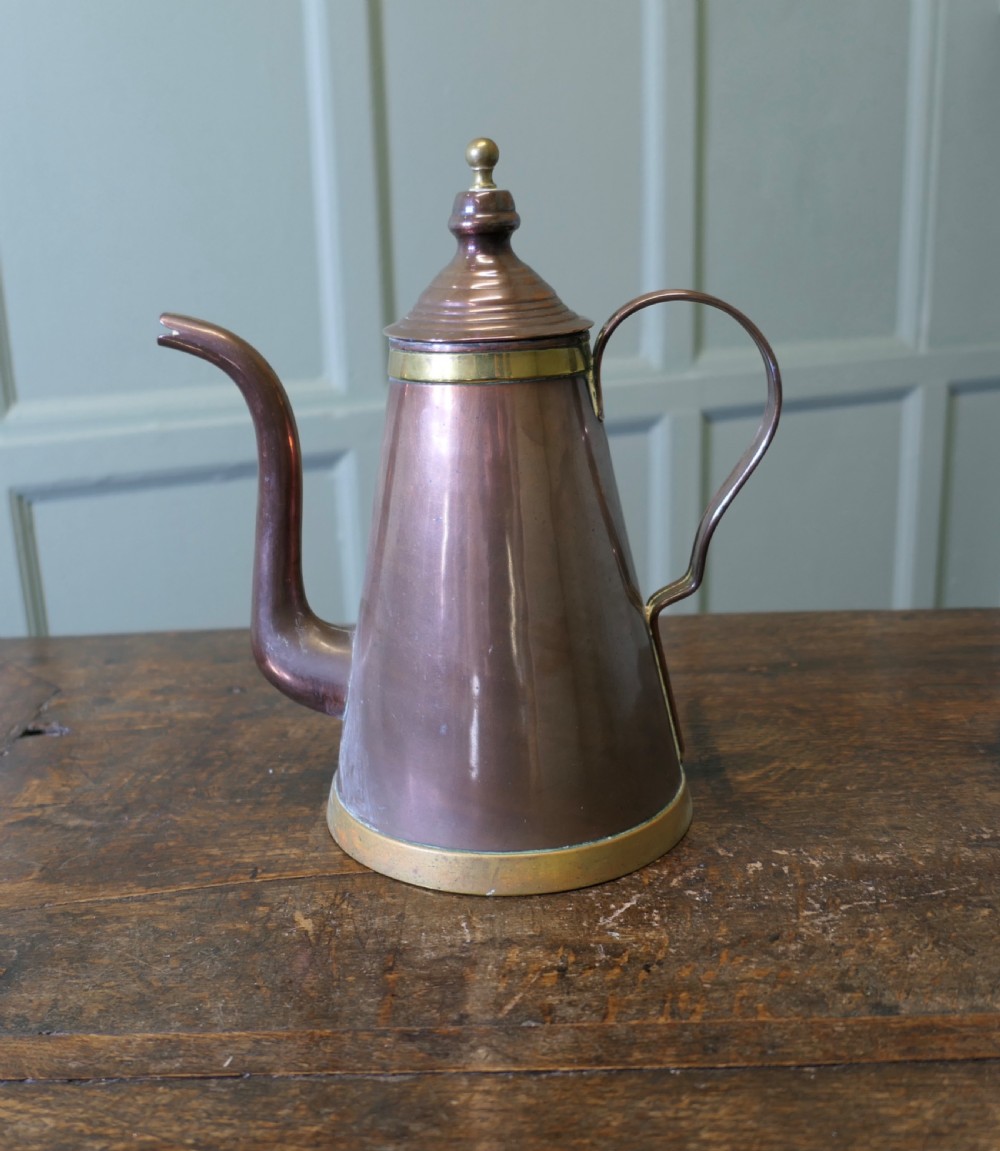 An Arts and Crafts copper coffee or chocolate pot with a conical body, brass collar, arched spout, strap handle, and finial lid. View II