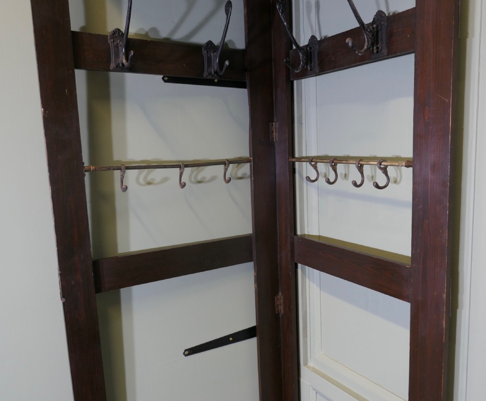 A tall, dark wooden corner coat stand featuring a hinged open framework, top shelf, cast iron coat hooks, and a brass hanging rail. View V