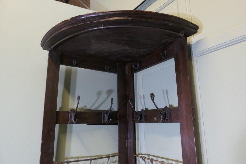 A tall, dark wooden corner coat stand featuring a hinged open framework, top shelf, cast iron coat hooks, and a brass hanging rail. View X