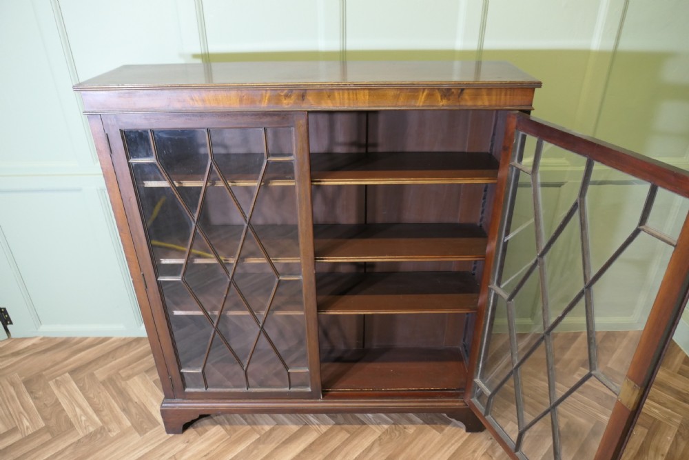 An Edwardian mahogany bookcase featuring two astragal glazed glass doors with geometric lattice patterns, internal wooden shelves, and bracket feet. View VI