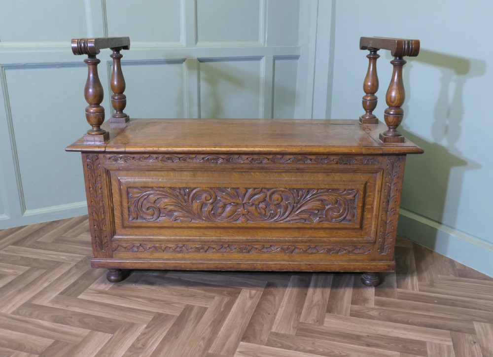 Carved golden oak storage coffer hall seat featuring a central Green Man mask relief carving and raised turned baluster arm supports.