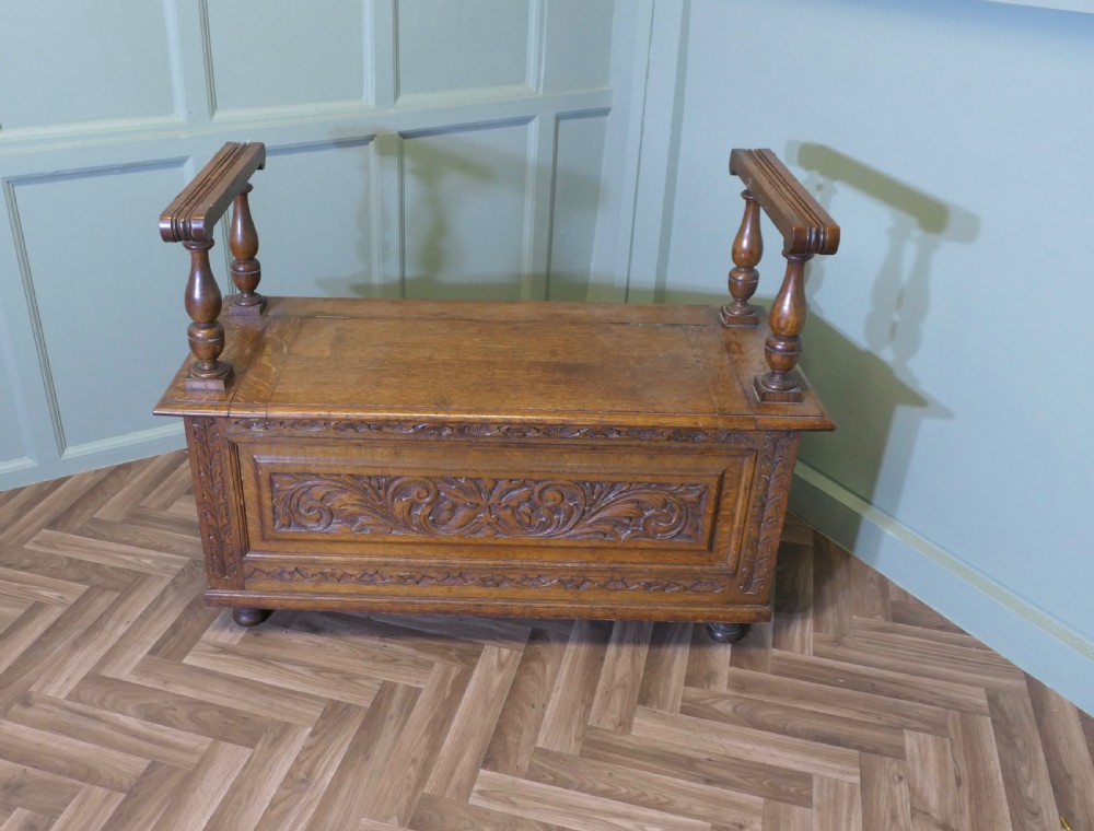 Carved golden oak storage coffer hall seat featuring a central Green Man mask relief carving and raised turned baluster arm supports. View II