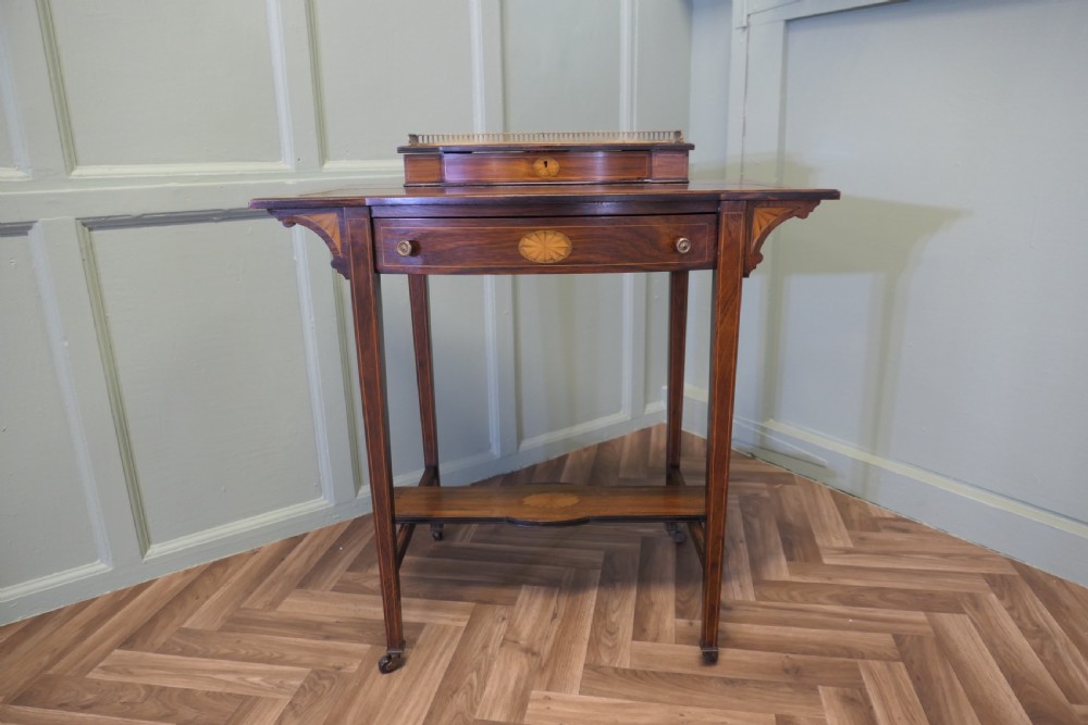 An Edwardian satin walnut ladies writing desk featuring a top superstructure with brass gallery, single drawer with marquetry oval inlay, tapered square legs, and an undertier shelf on brass castors.