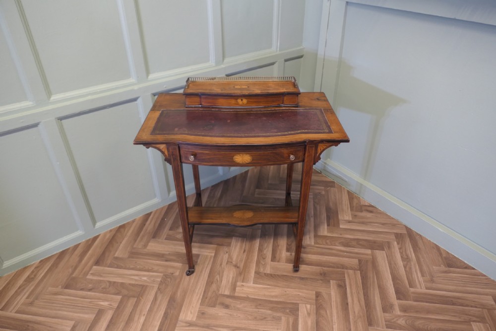 An Edwardian satin walnut ladies writing desk featuring a top superstructure with brass gallery, single drawer with marquetry oval inlay, tapered square legs, and an undertier shelf on brass castors. View II