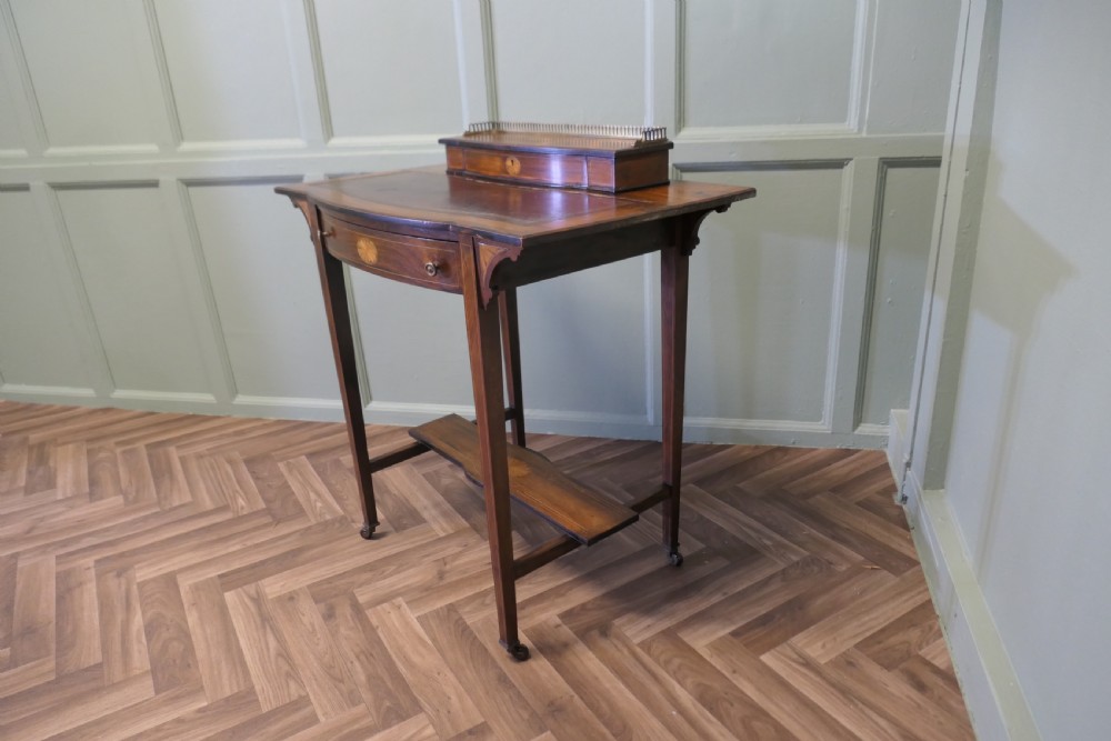 An Edwardian satin walnut ladies writing desk featuring a top superstructure with brass gallery, single drawer with marquetry oval inlay, tapered square legs, and an undertier shelf on brass castors. View III
