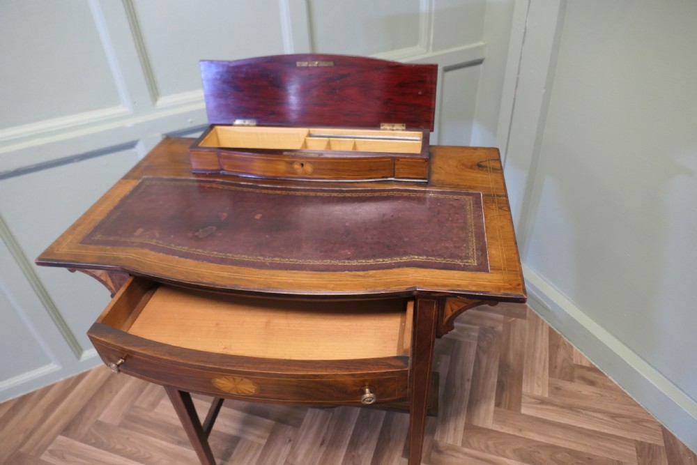 An Edwardian satin walnut ladies writing desk featuring a top superstructure with brass gallery, single drawer with marquetry oval inlay, tapered square legs, and an undertier shelf on brass castors. View VI