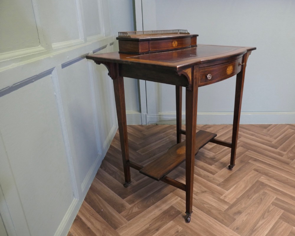 An Edwardian satin walnut ladies writing desk featuring a top superstructure with brass gallery, single drawer with marquetry oval inlay, tapered square legs, and an undertier shelf on brass castors. View VIII