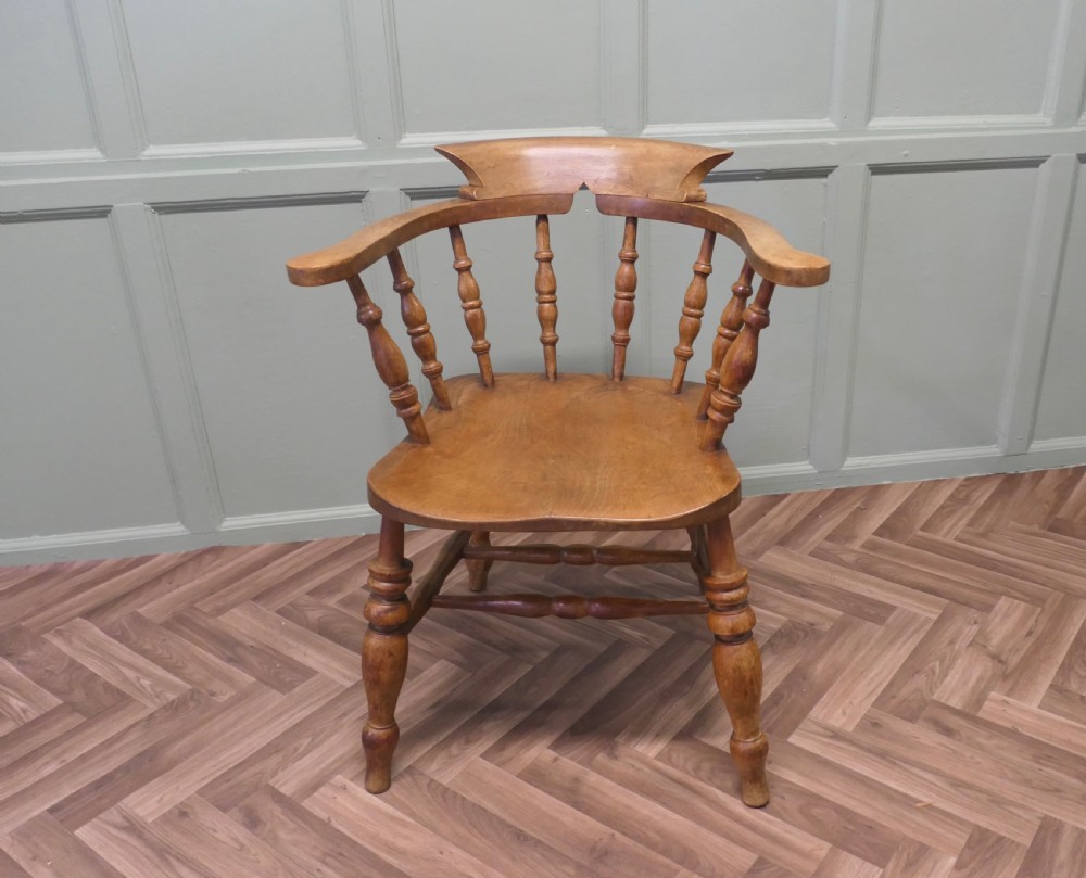 A 19th-century wooden smokers bow armchair featuring a curved crest rail, turned baluster spindles, a carved saddle seat, and turned legs.