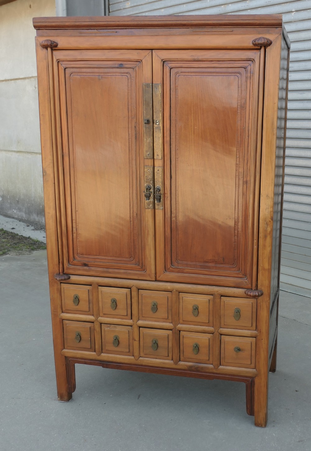 A tall warm brown teak cupboard featuring two upper panelled doors with brass hardware above ten small drawers arranged in two rows. View II