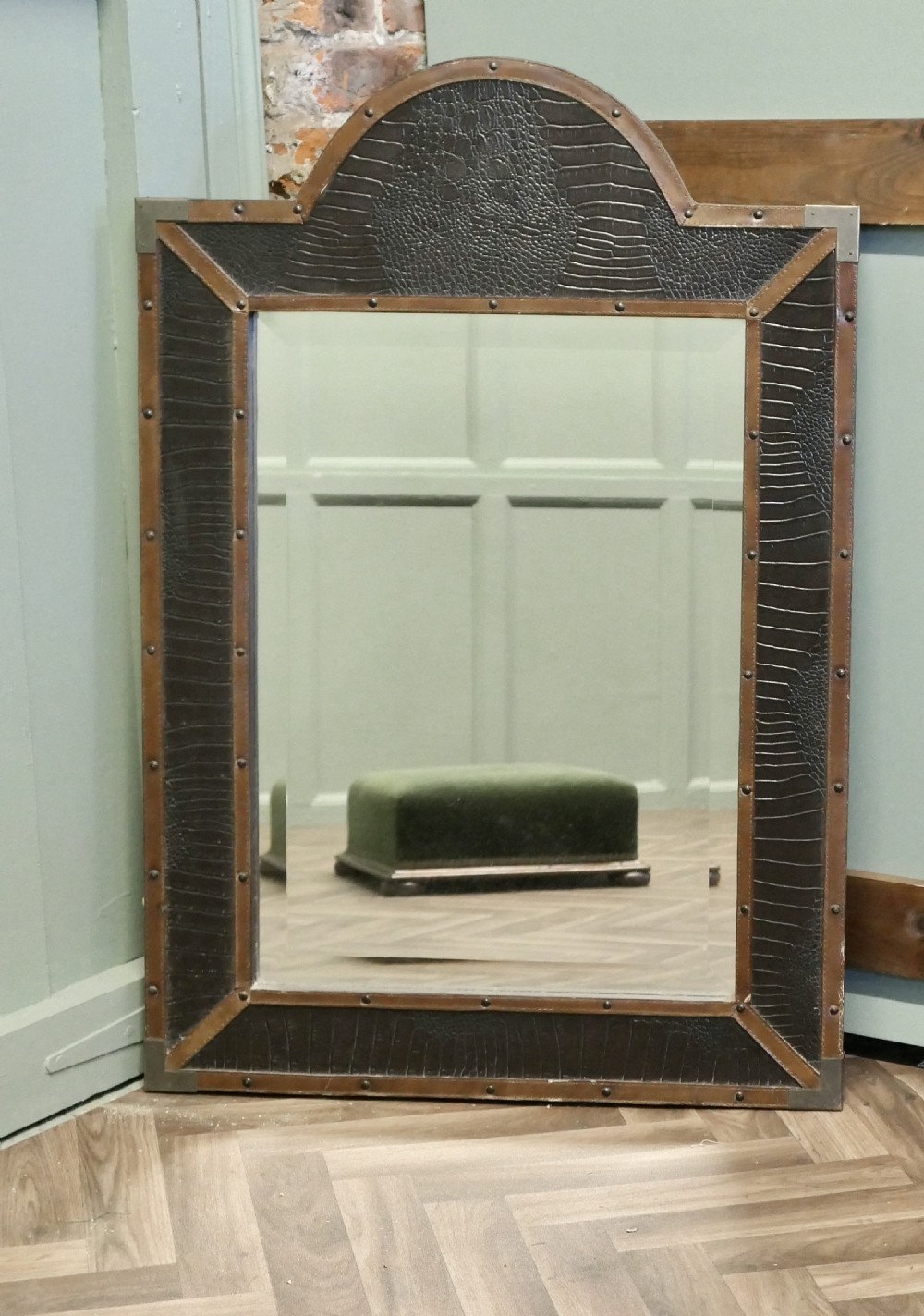 An Art Deco wall mirror featuring a dark crocodile-embossed leather frame with tan leather borders, brass stud details, and an arched crest.