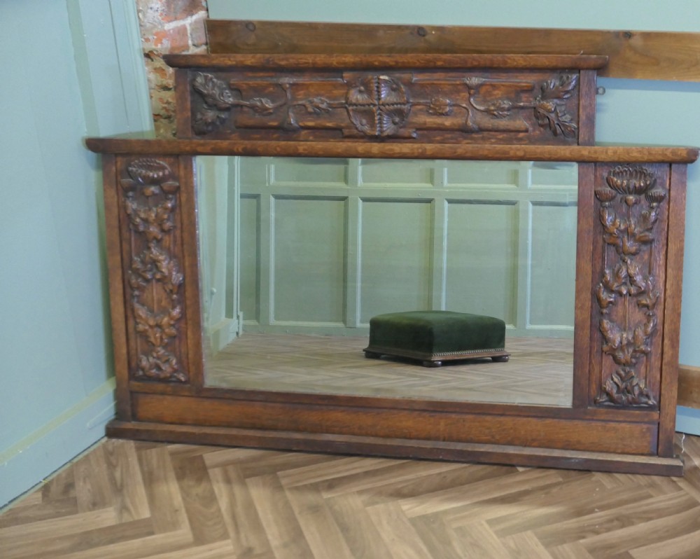 A large Arts and Crafts oak overmantel mirror featuring detailed relief carvings of thistles and stylized leaves around a rectangular glass mirror.