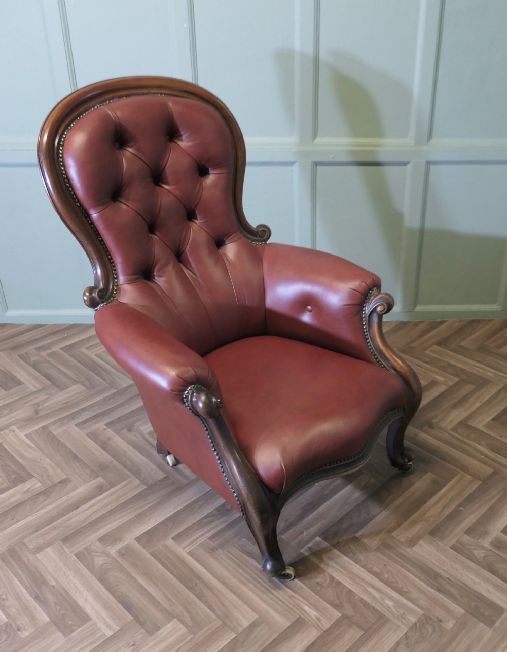 Victorian button-back salon armchair upholstered in burgundy leather with a carved dark wood frame and brass castors.
