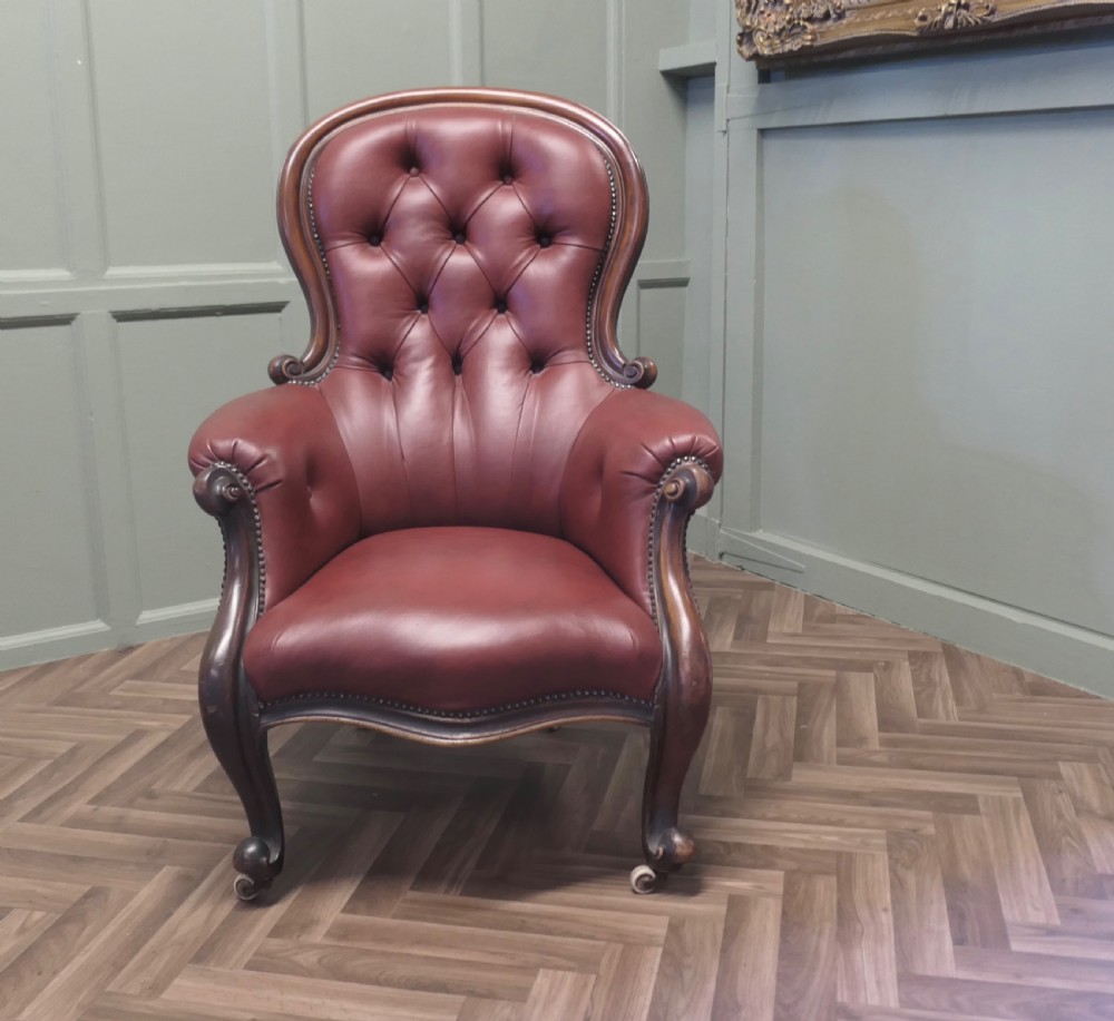 Victorian button-back salon armchair upholstered in burgundy leather with a carved dark wood frame and brass castors. View II