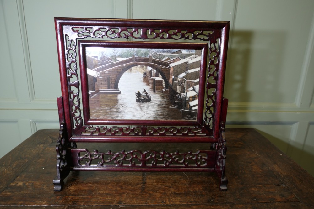 A detailed Chinese silk embroidery depicting a canal and boat scene set inside a dark carved hardwood table screen.