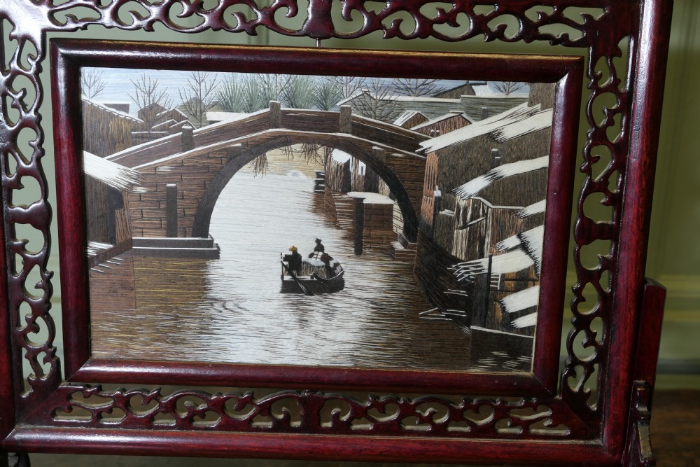 A detailed Chinese silk embroidery depicting a canal and boat scene set inside a dark carved hardwood table screen. View II