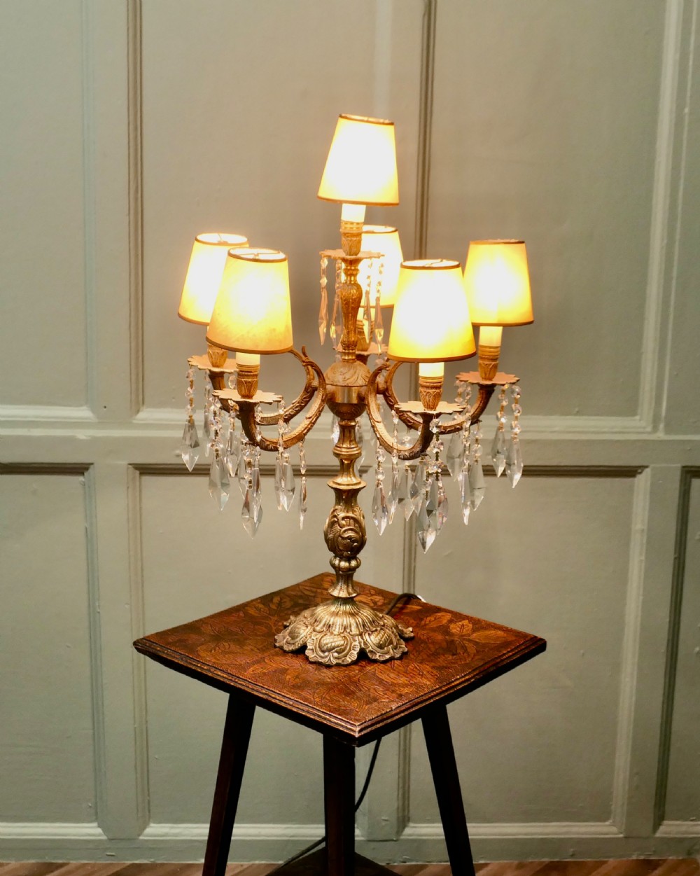 An ornate French brass six-arm girandole table lamp featuring hanging crystal prisms and small cream lampshades.