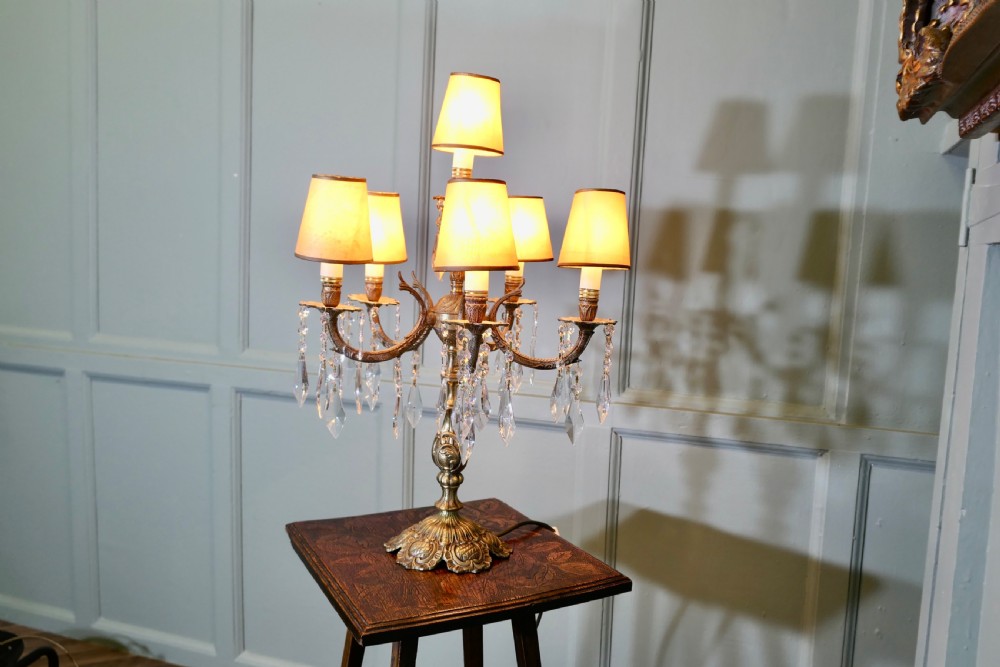 An ornate French brass six-arm girandole table lamp featuring hanging crystal prisms and small cream lampshades. View III