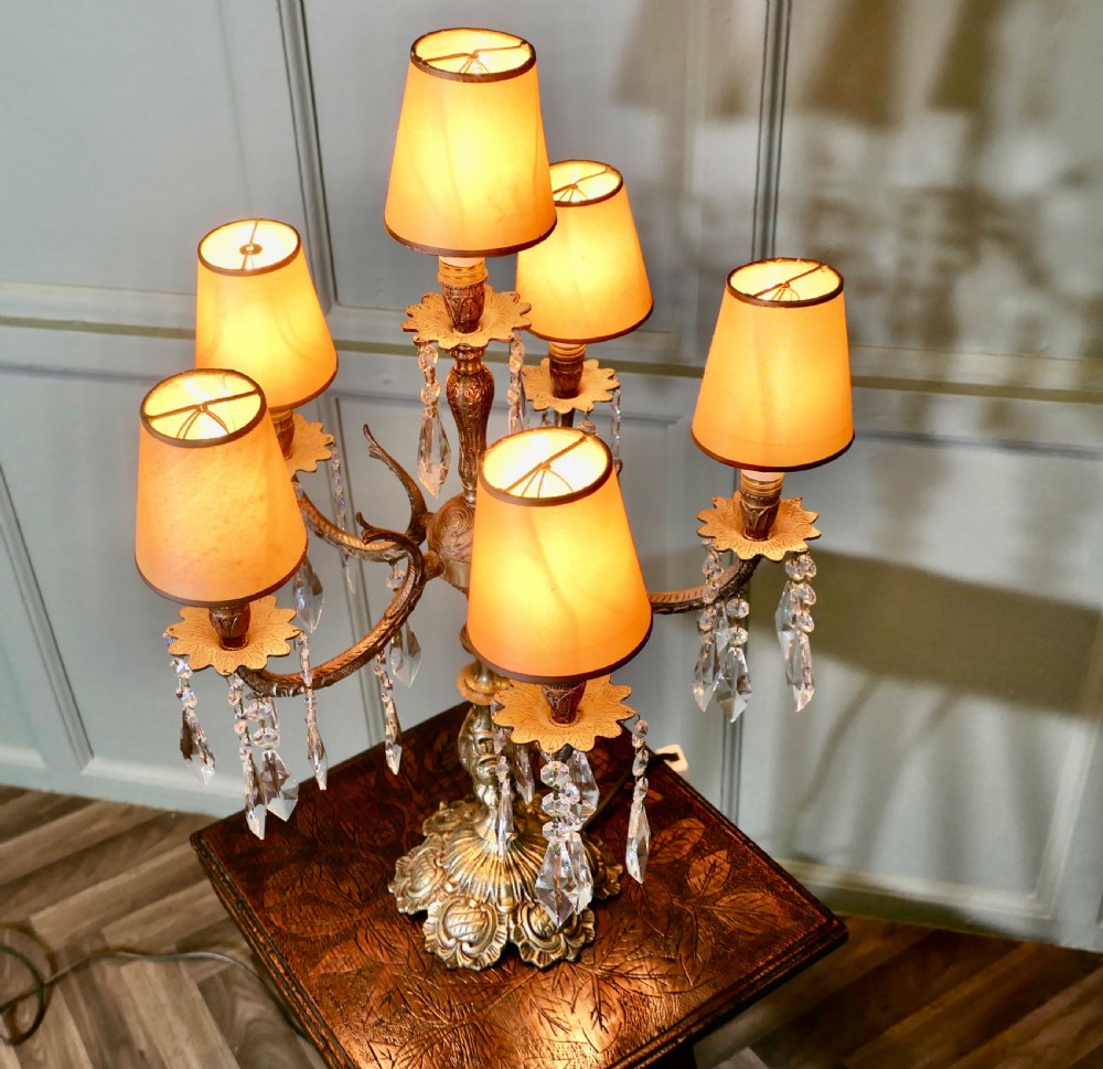 An ornate French brass six-arm girandole table lamp featuring hanging crystal prisms and small cream lampshades. View IV