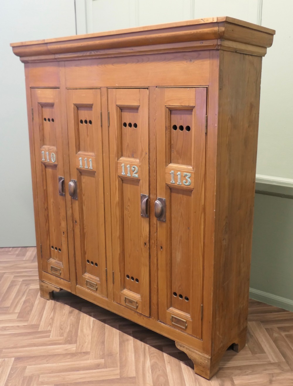 A four-door golden pine sports locker featuring numbered metal tags, circular ventilation holes, and brass fittings. View V