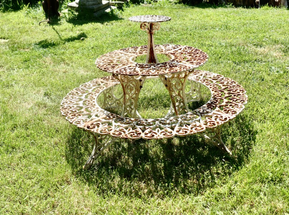 A circular three-tiered cast iron plant stand featuring intricate openwork scrollwork with weathered white paint and visible surface rust. View II