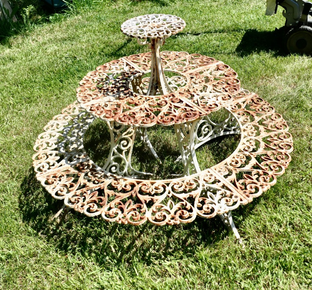 A circular three-tiered cast iron plant stand featuring intricate openwork scrollwork with weathered white paint and visible surface rust. View IV