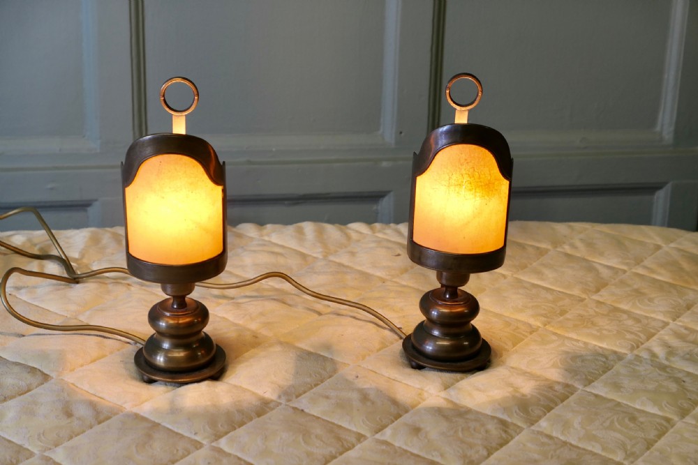 A pair of turned brass bedside lamps with railway lantern styled cowls, ring finials, and illuminated amber parchment shades.