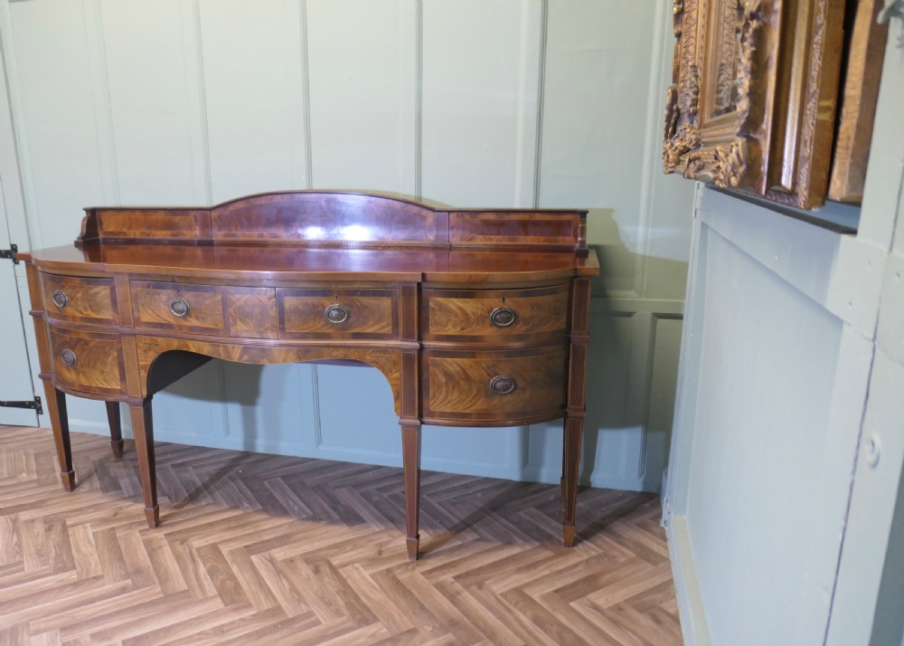 A 19th-century mahogany bow-fronted sideboard featuring flame veneer drawer fronts, brass drop ring handles, a raised upstand, and tapered square legs.