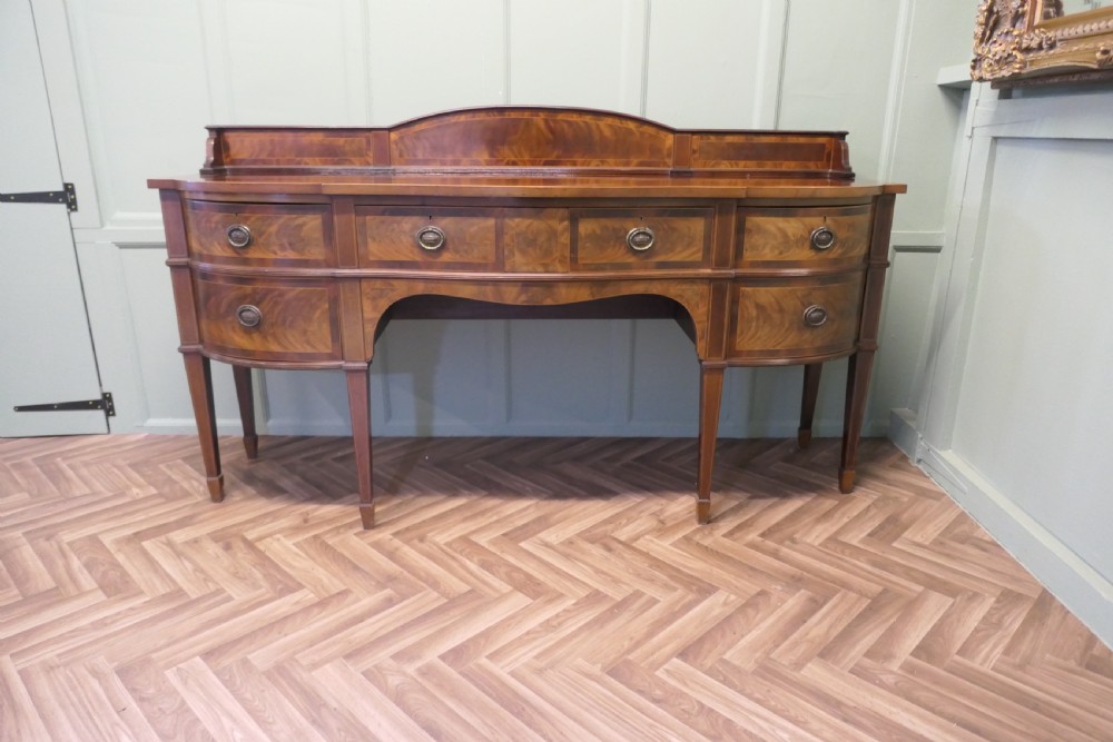 A 19th-century mahogany bow-fronted sideboard featuring flame veneer drawer fronts, brass drop ring handles, a raised upstand, and tapered square legs. View V