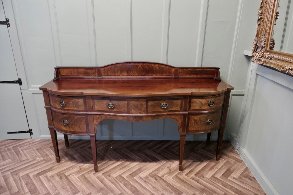 A 19th-century mahogany bow-fronted sideboard featuring flame veneer drawer fronts, brass drop ring handles, a raised upstand, and tapered square legs. View VI
