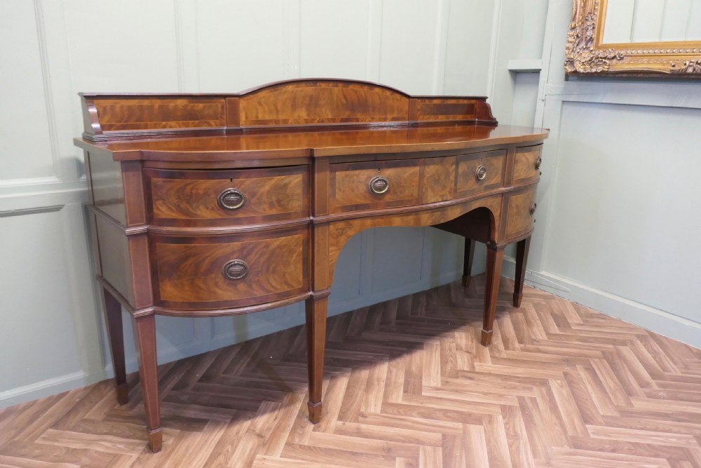 A 19th-century mahogany bow-fronted sideboard featuring flame veneer drawer fronts, brass drop ring handles, a raised upstand, and tapered square legs. View VII