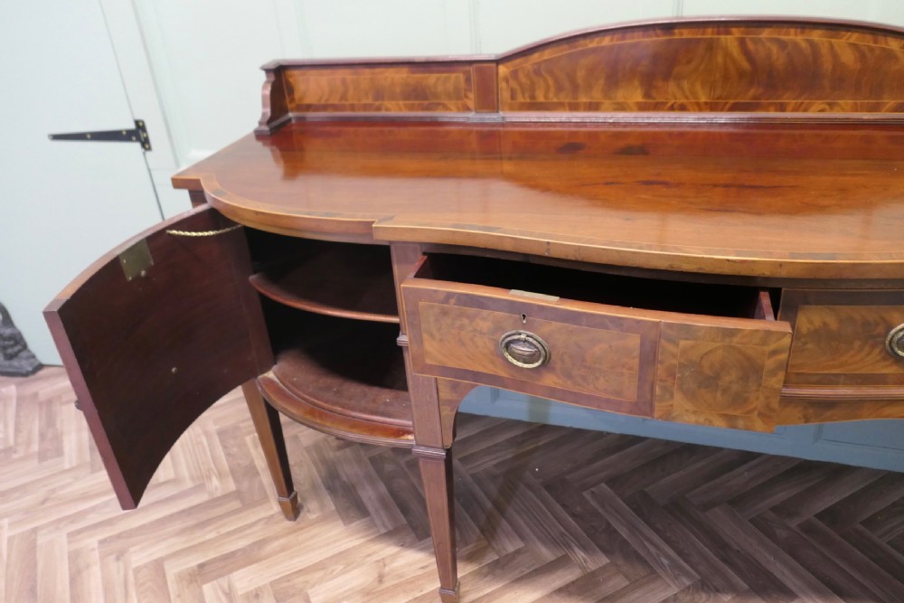 A 19th-century mahogany bow-fronted sideboard featuring flame veneer drawer fronts, brass drop ring handles, a raised upstand, and tapered square legs. View VIII