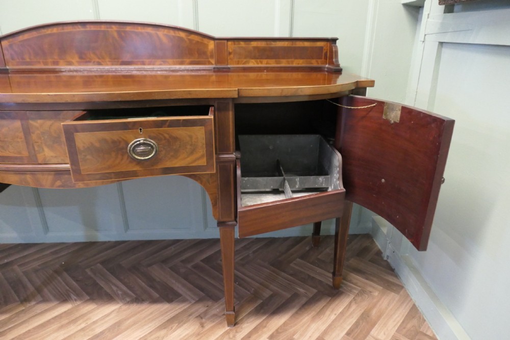 A 19th-century mahogany bow-fronted sideboard featuring flame veneer drawer fronts, brass drop ring handles, a raised upstand, and tapered square legs. View IX