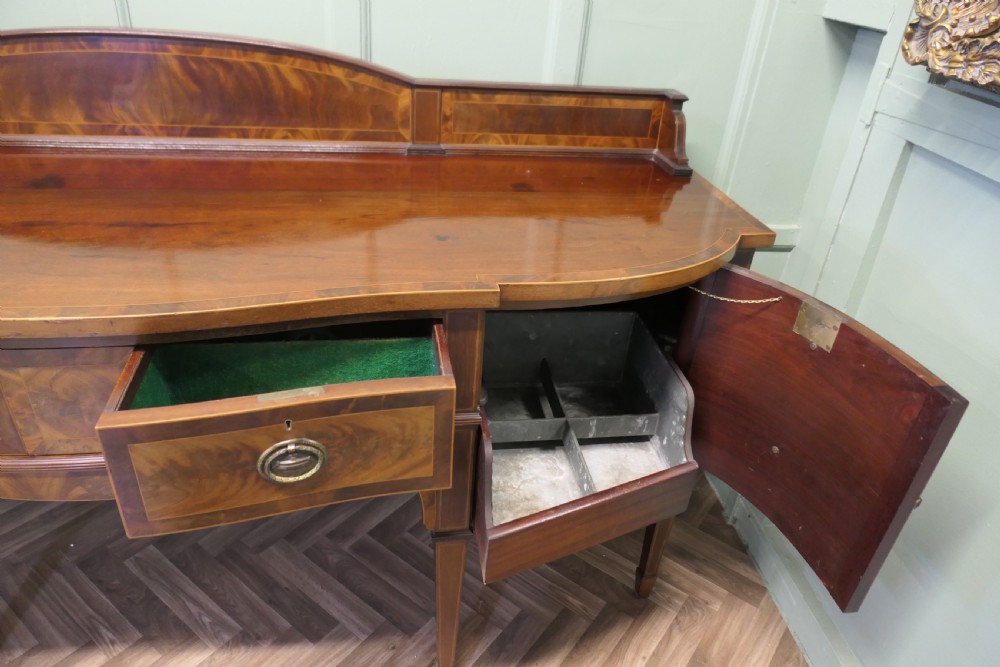 A 19th-century mahogany bow-fronted sideboard featuring flame veneer drawer fronts, brass drop ring handles, a raised upstand, and tapered square legs. View X