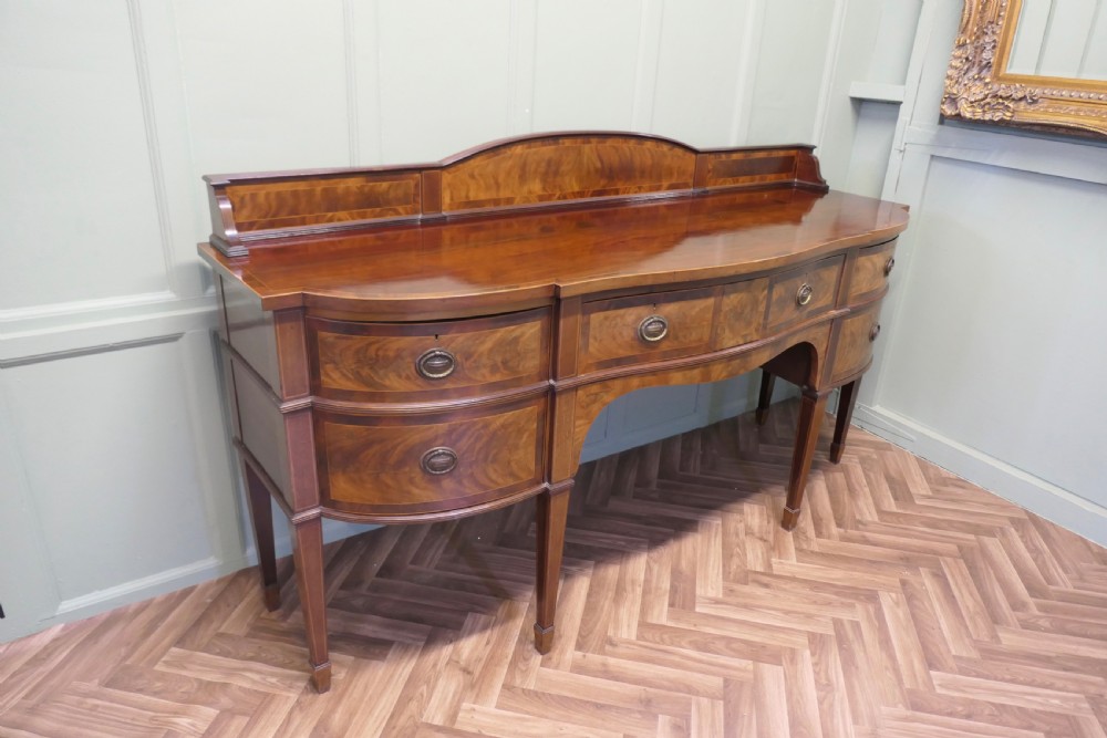 A 19th-century mahogany bow-fronted sideboard featuring flame veneer drawer fronts, brass drop ring handles, a raised upstand, and tapered square legs. View XI