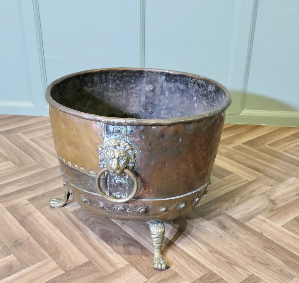 A large, circular antique copper log cauldron featuring hand-riveted construction, cast brass lion mask handles, and claw feet. View III