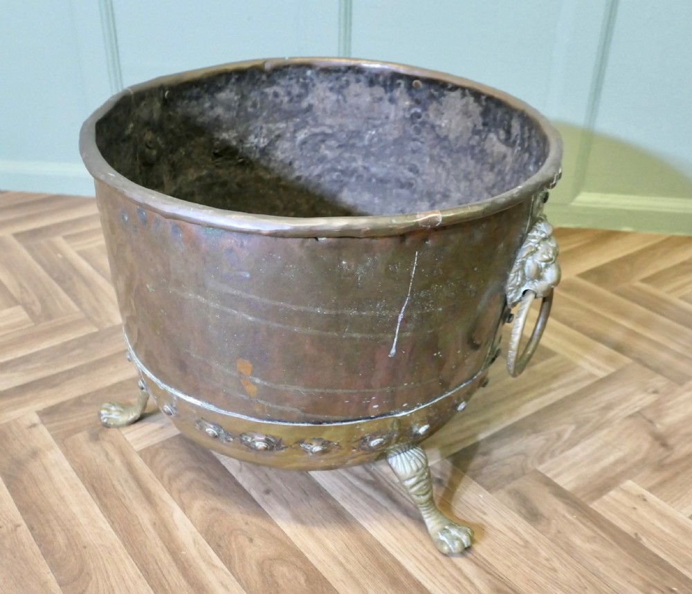 A large, circular antique copper log cauldron featuring hand-riveted construction, cast brass lion mask handles, and claw feet. View IV