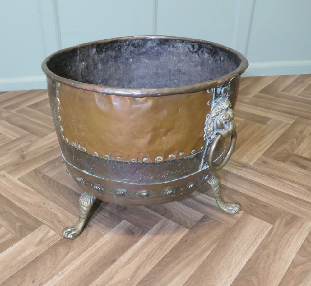 A large, circular antique copper log cauldron featuring hand-riveted construction, cast brass lion mask handles, and claw feet. View V