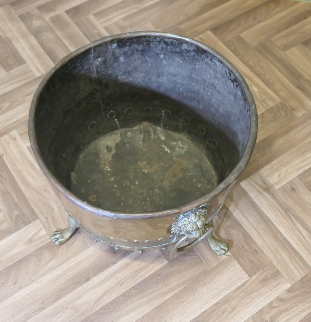 A large, circular antique copper log cauldron featuring hand-riveted construction, cast brass lion mask handles, and claw feet. View VI