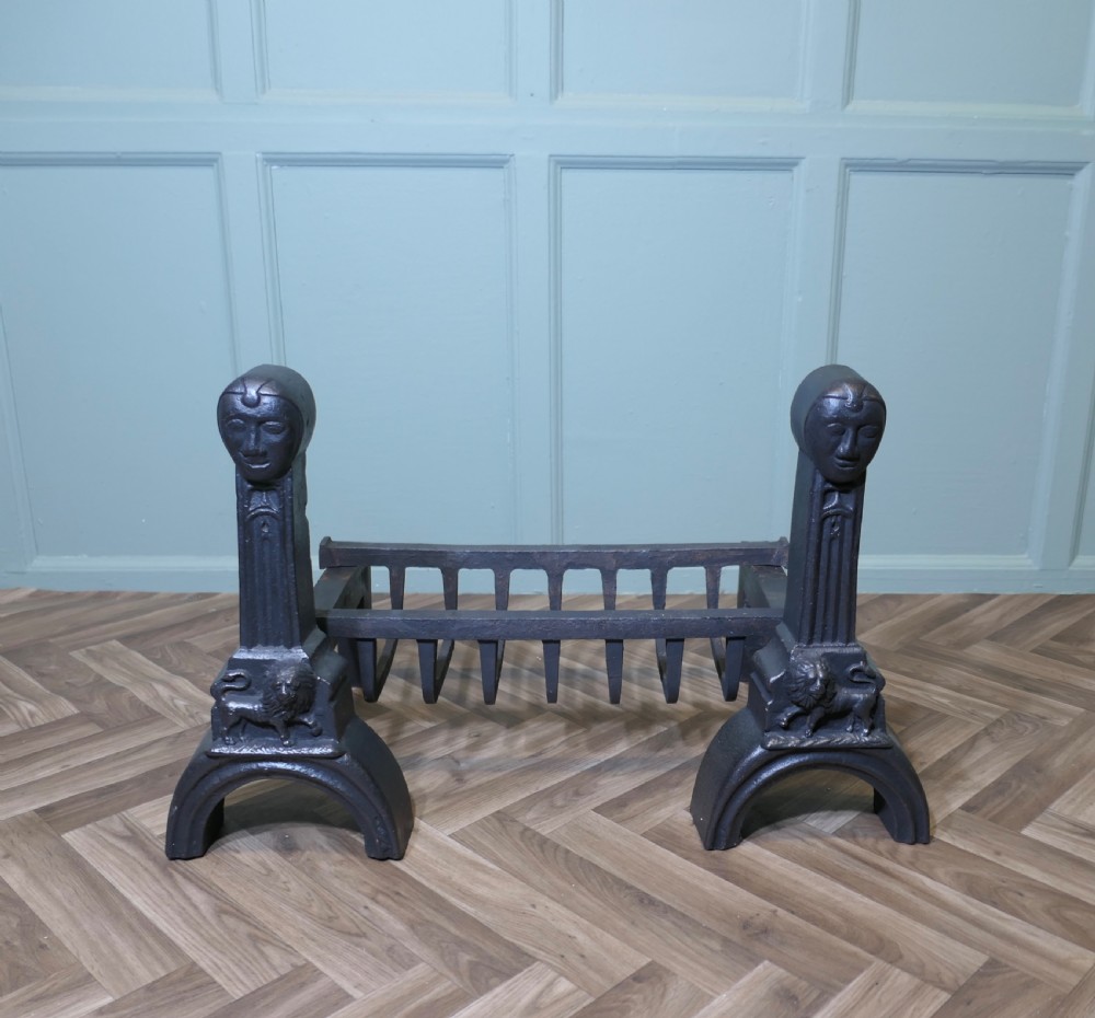 A heavy black cast iron inglenook fire grate supported by two ornate firedogs featuring sculpted relief heads and lion bases.