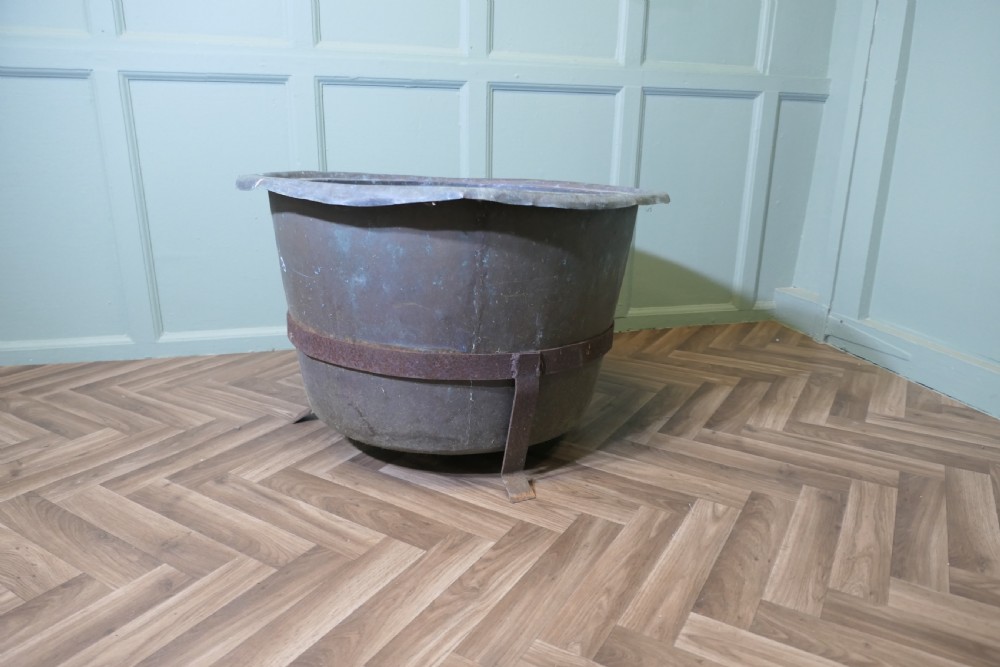 A large round patinated copper vessel with a rolled rim resting inside a dark wrought iron stand.